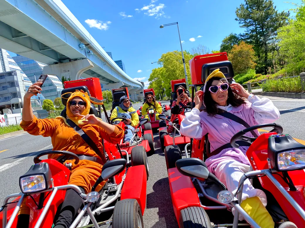 Group of people in bright animal costumes riding red go-karts on a sunny street, posing for photos and selfies as they ride in a line.