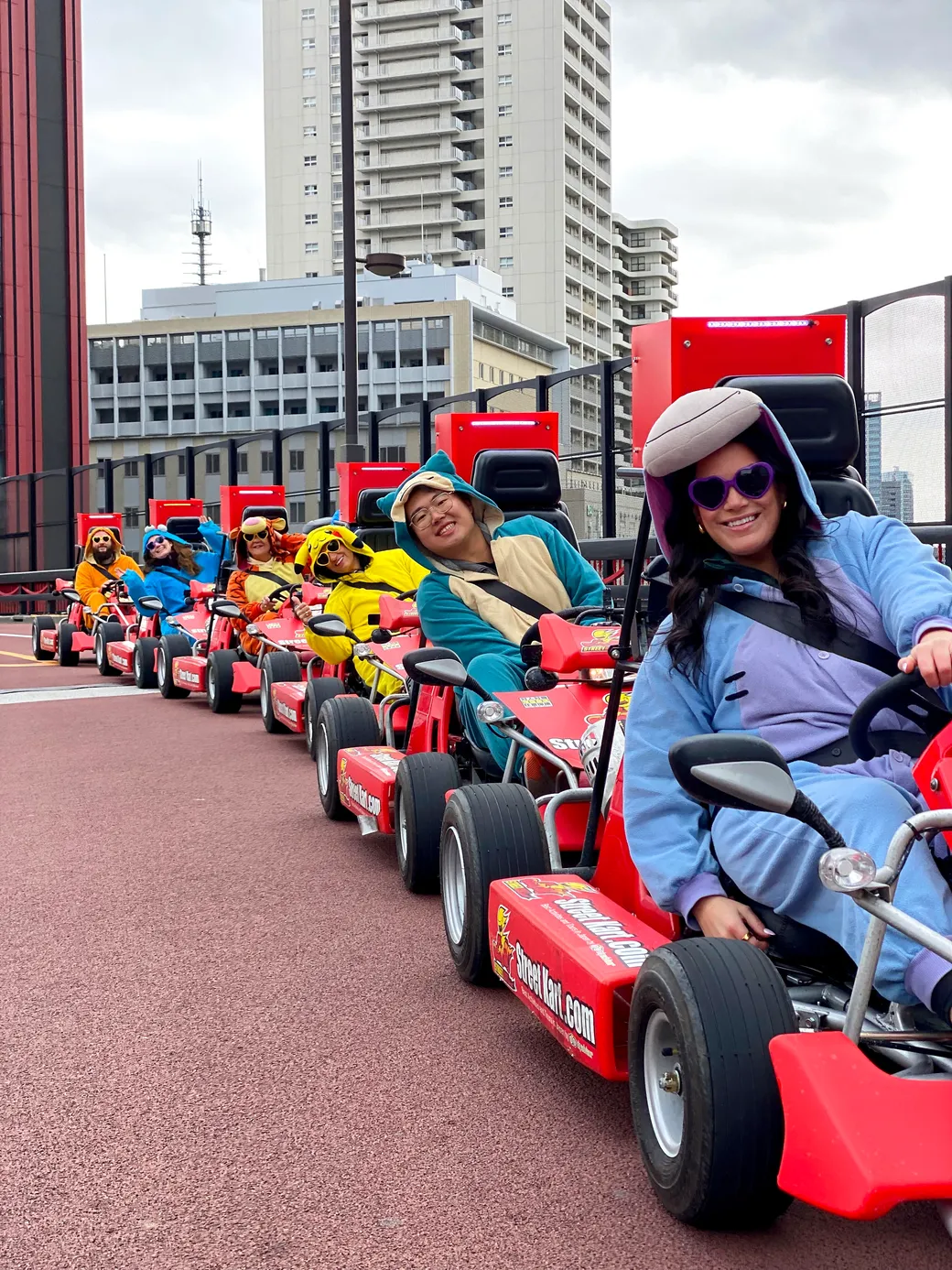 Group of people driving bright red go-karts in a city outdoor track, wearing colorful hoodies and helmets with smiles.