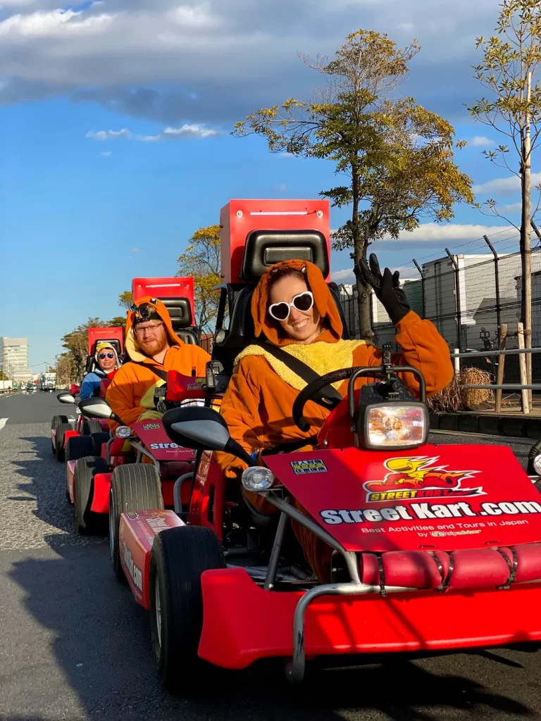 IMG_3600 - StreetKart Person in an orange mascot outfit with heart-shaped sunglasses drives a red Street Kart go-kart, waving at the camera; others in orange costumes follow in line behind.