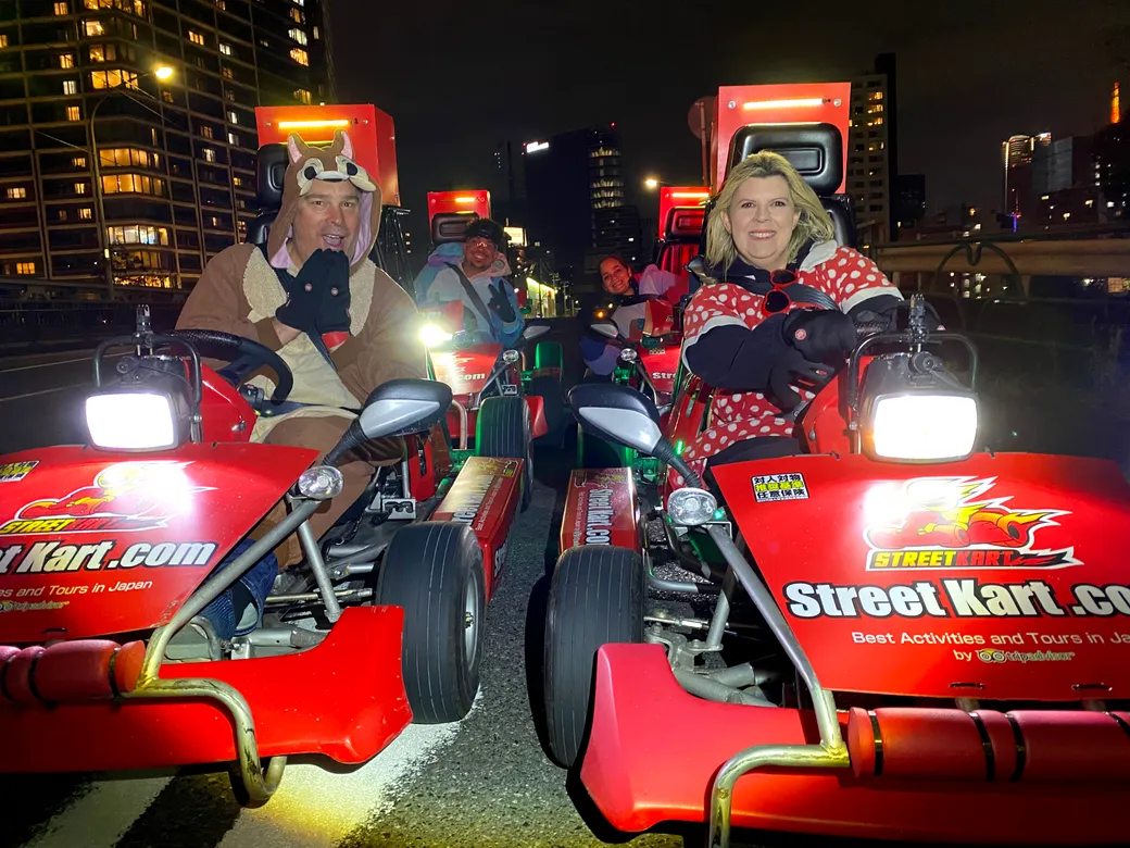 Group of people in red street go-karts at night, some in animal and polka-dot costumes, smiling in a city street race setting with tall buildings in the background.